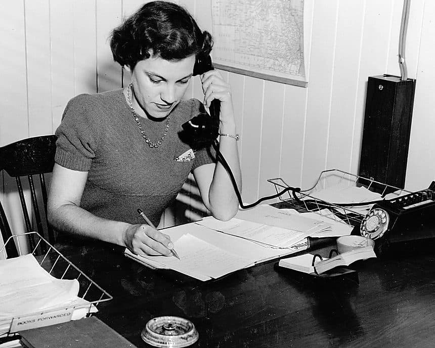 Archival photograph of a clerk taking notes at a desk with a telephone and typewriter.